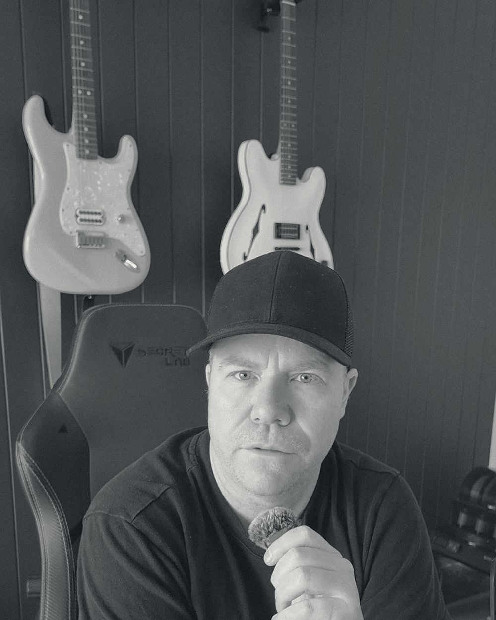 Paul Warren in the Melbourne studio, arms crossed, tattooed forearm, cap on, guitars on the wall above him. Black and white.