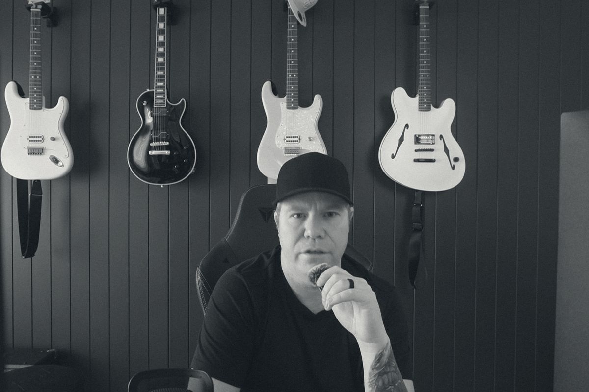 Paul Warren at the microphone in the Melbourne studio, black cap and black t-shirt, four guitars on the wall behind him. Black and white.